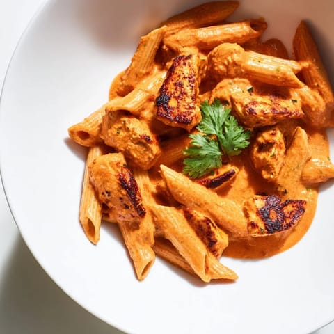 A close-up of golden-brown chicken tikka pieces nestled in a rich, tomato-cream sauce over al dente penne pasta.